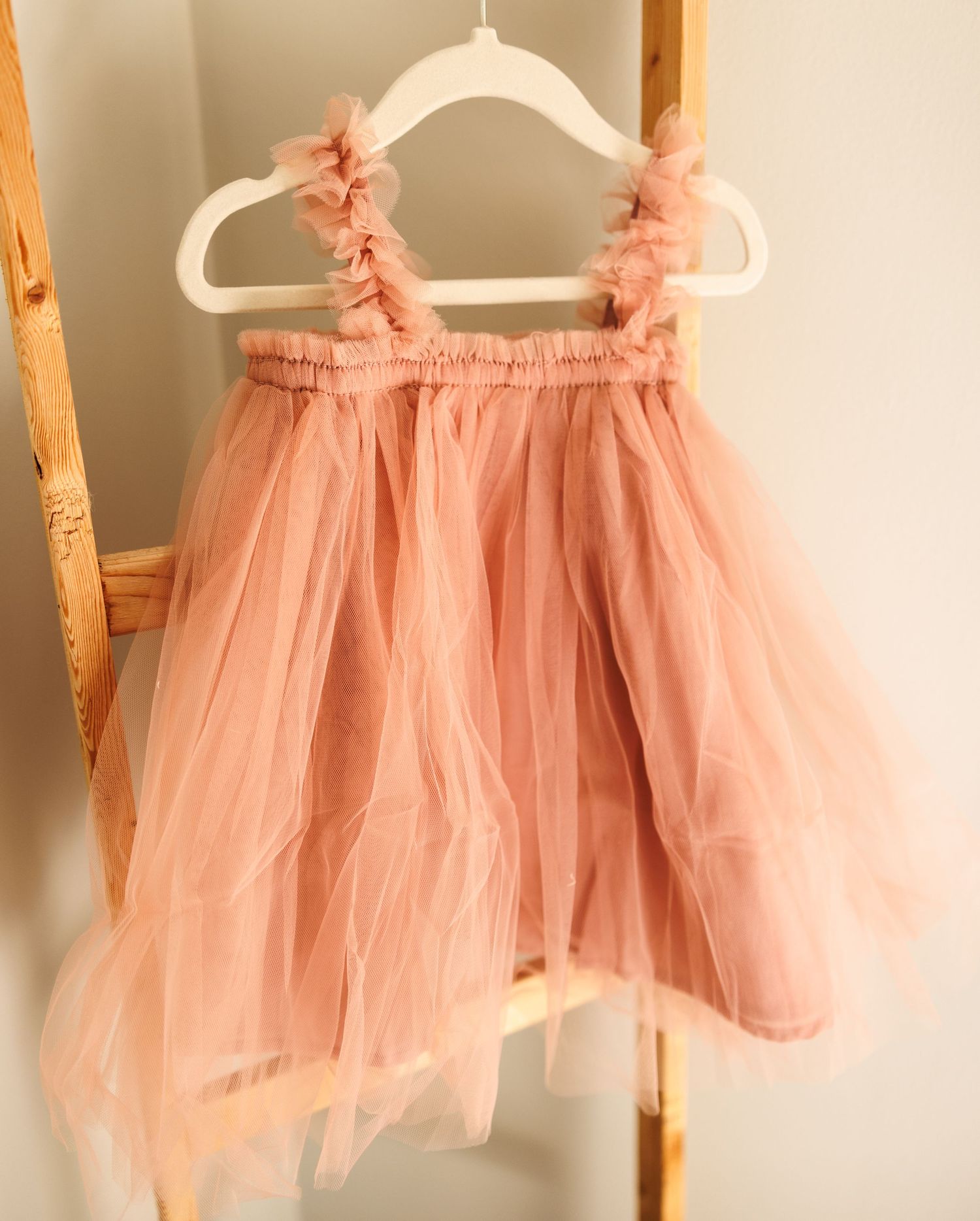 A delicate pink tulle dress with ruffle straps hangs on a white plastic hanger against a beige wall.