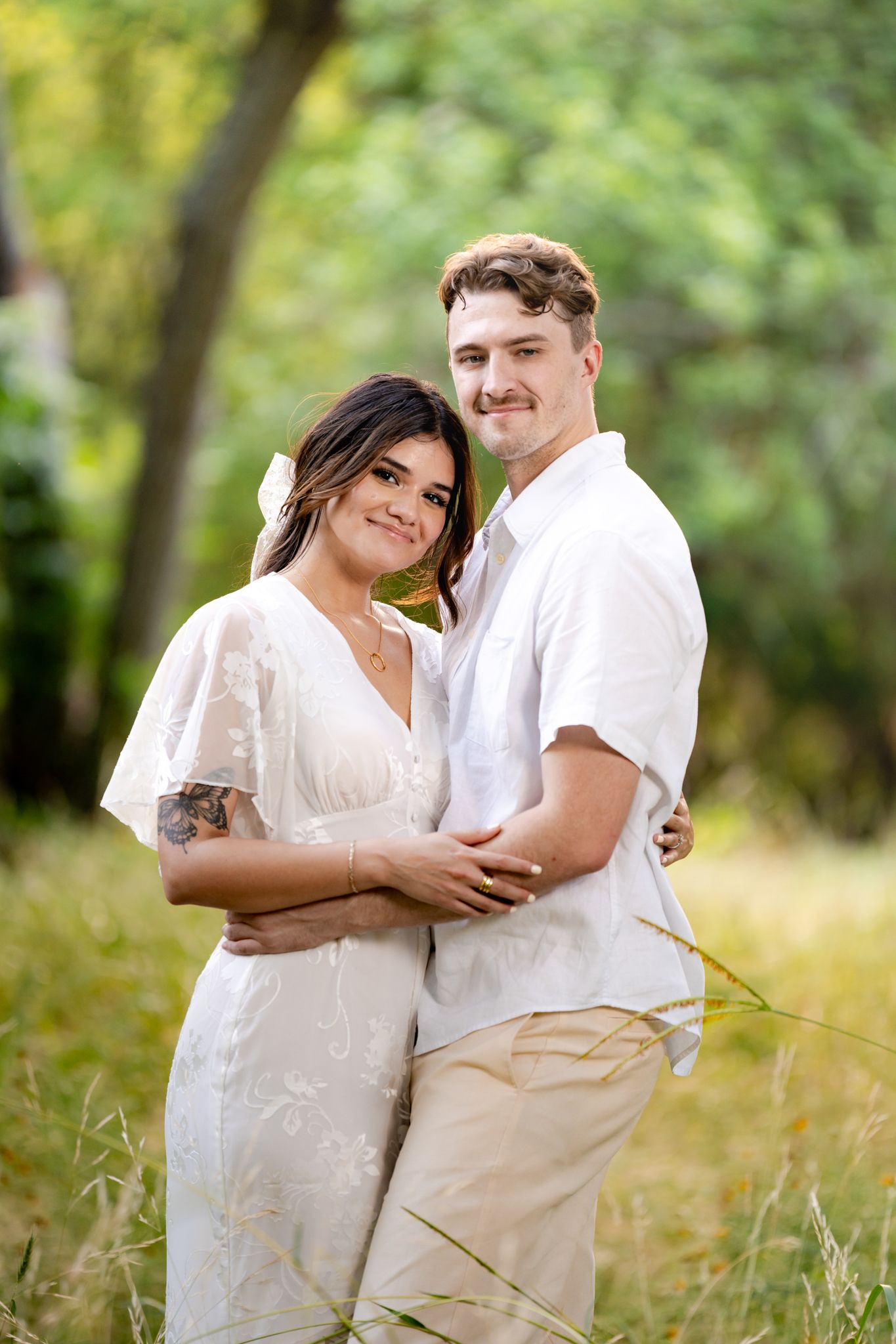 A couple shares an intimate moment outdoors in a lush green setting while wearing white and khaki clothing.