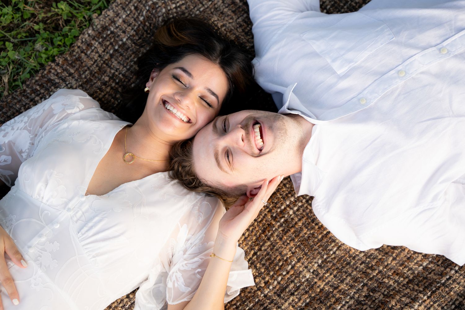 Two people lying on woven blanket outdoors with lively expressions of joy and laughter.