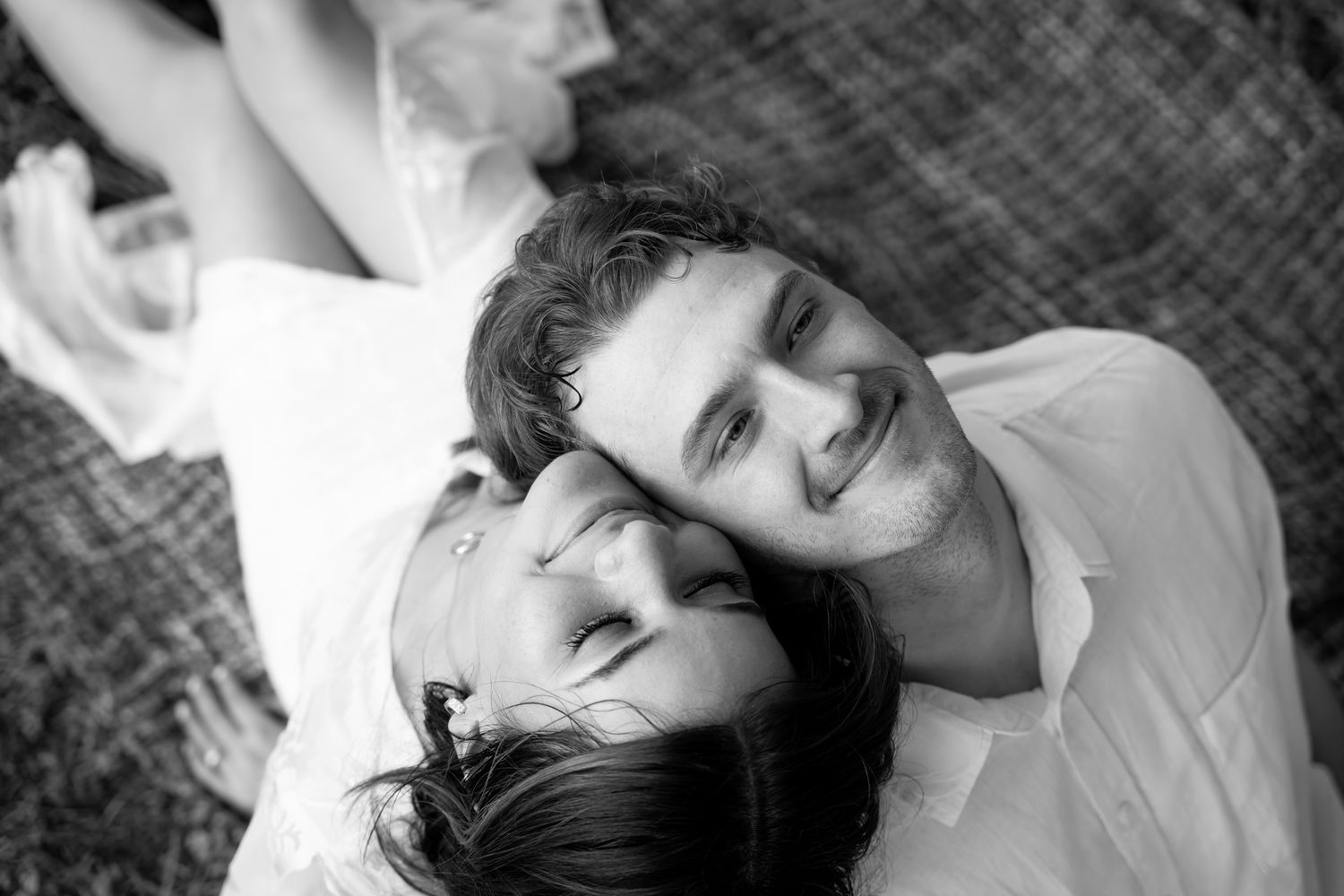 A black and white photograph shows two people lying close together and smiling while looking up at the camera.