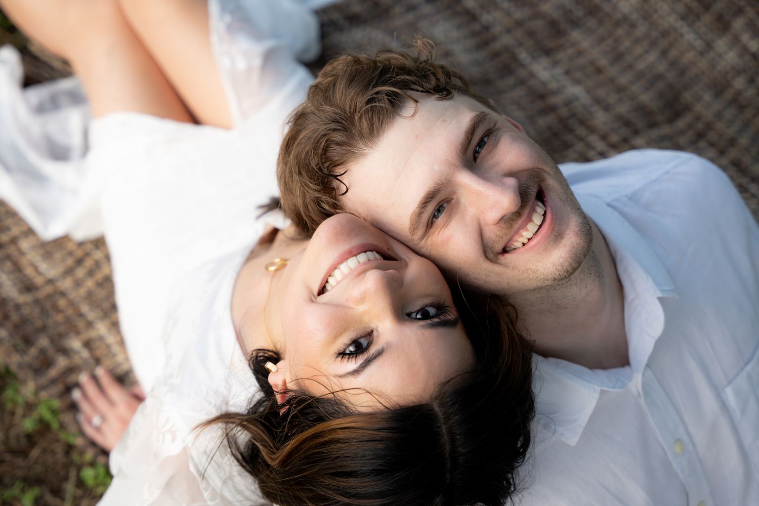 A couple wearing white clothes lying down together on a textured surface while smiling at the camera.