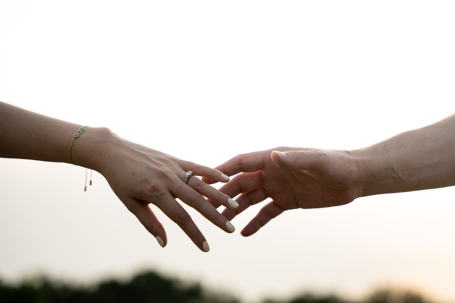 Two silhouetted hands reaching toward each other against a soft, bright background at sunset.