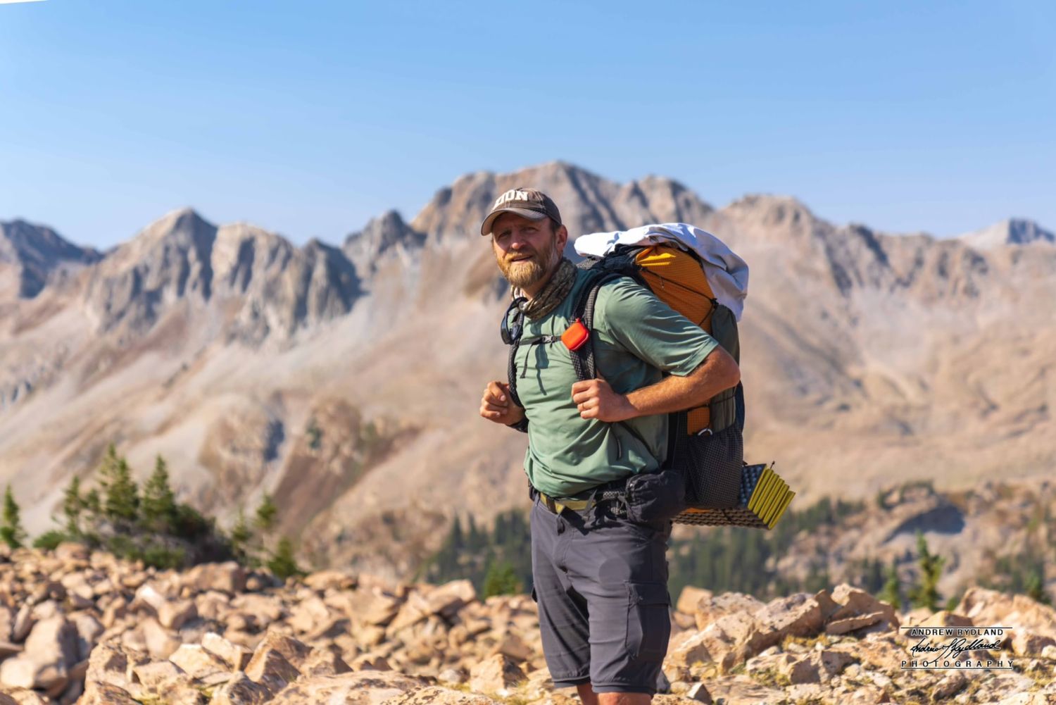 Hiker traverses rocky mountain terrain with dramatic peaks in background.