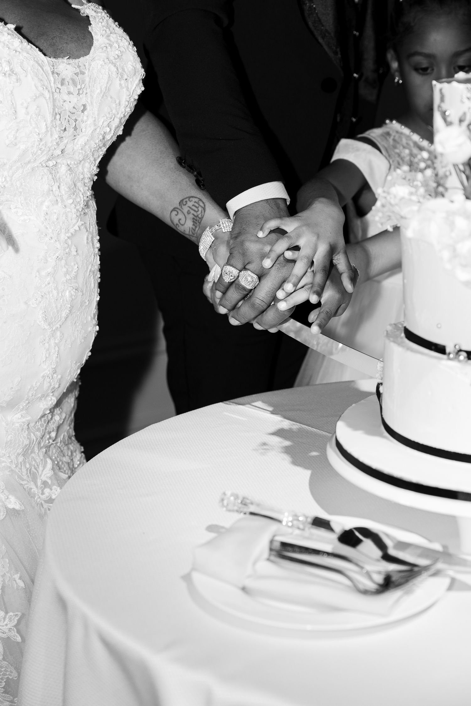 Black and white image of two hands cutting a tiered wedding cake together while wearing wedding attire.