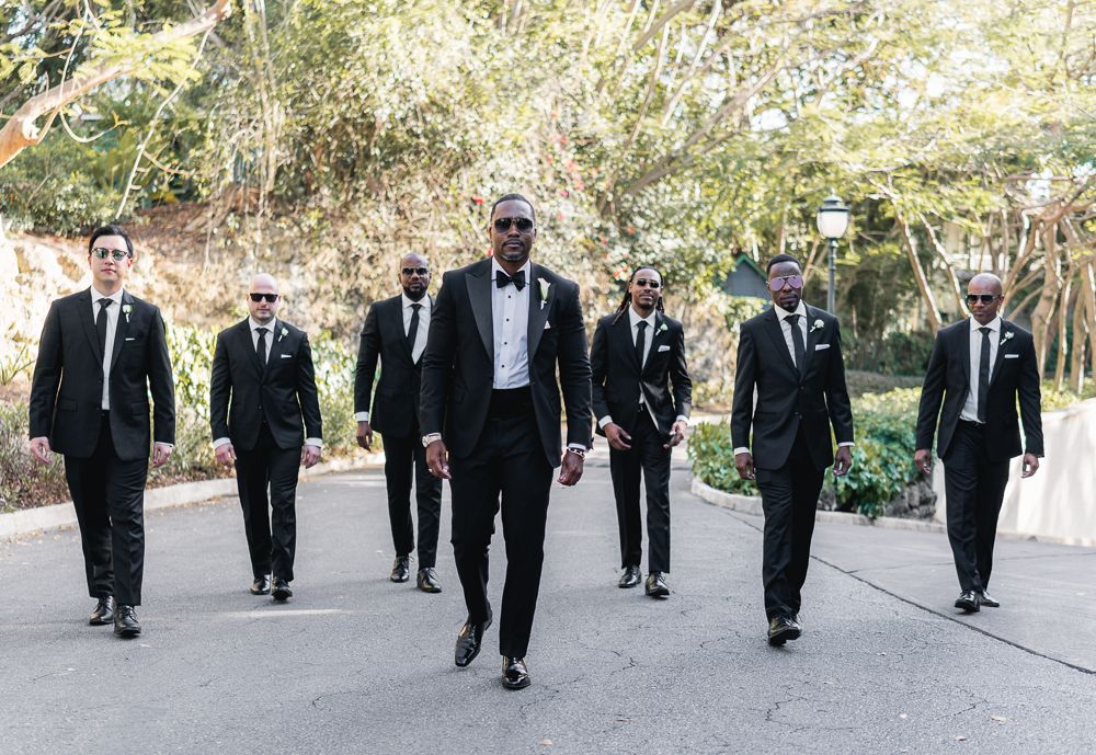 Groomsmen wearing black tuxedos and bowties walk together on a path surrounded by greenery.
