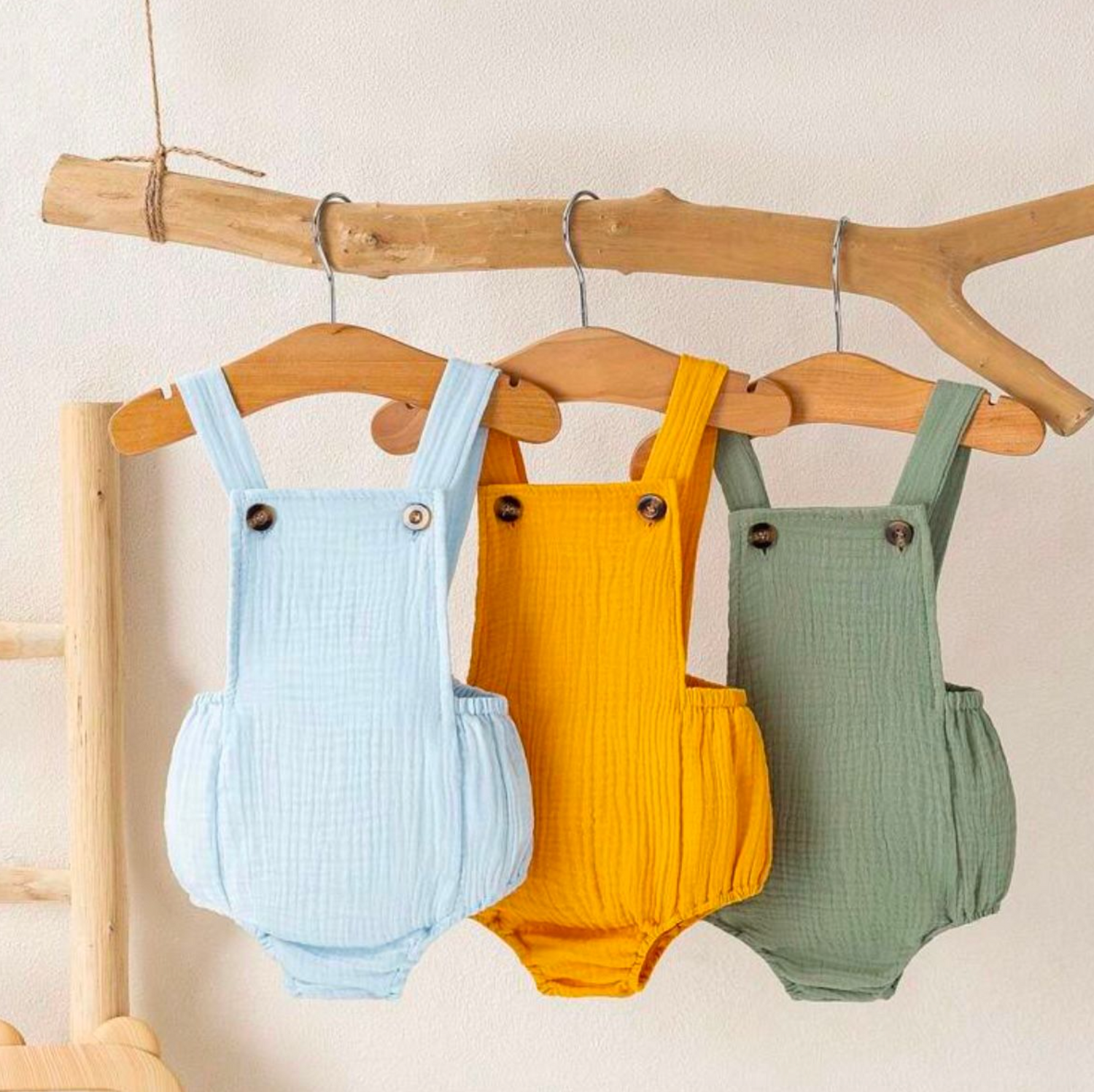 Baby rompers in light blue, mustard yellow and sage green displayed on rustic wooden branch.
