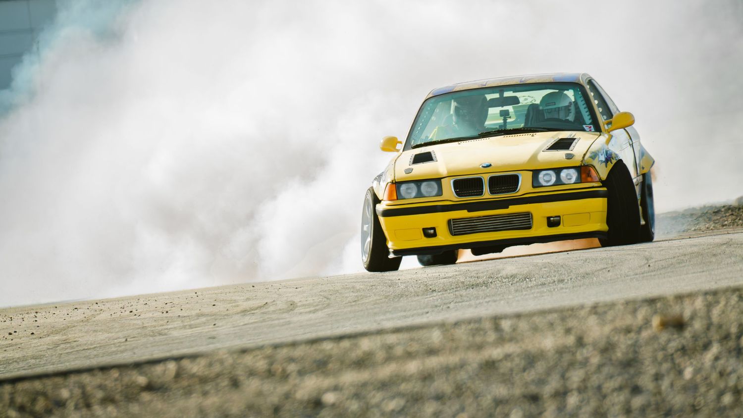 Yellow BMW M3 drifting on a mountain road creating a cloud of tire smoke.