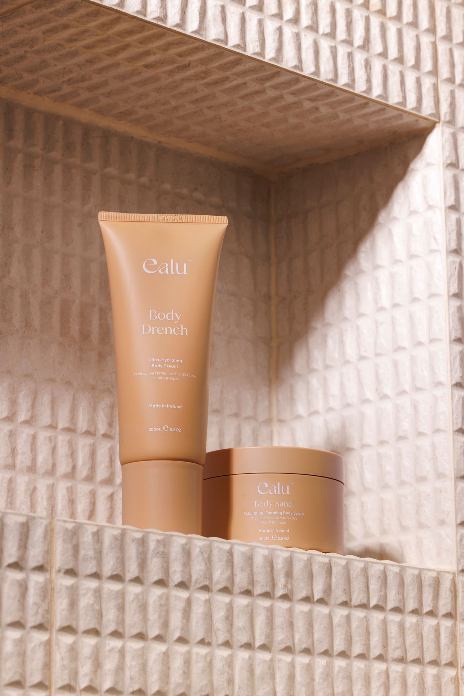 Two brown skincare product tubes displayed on a textured cream-colored shelf in a bathroom niche.