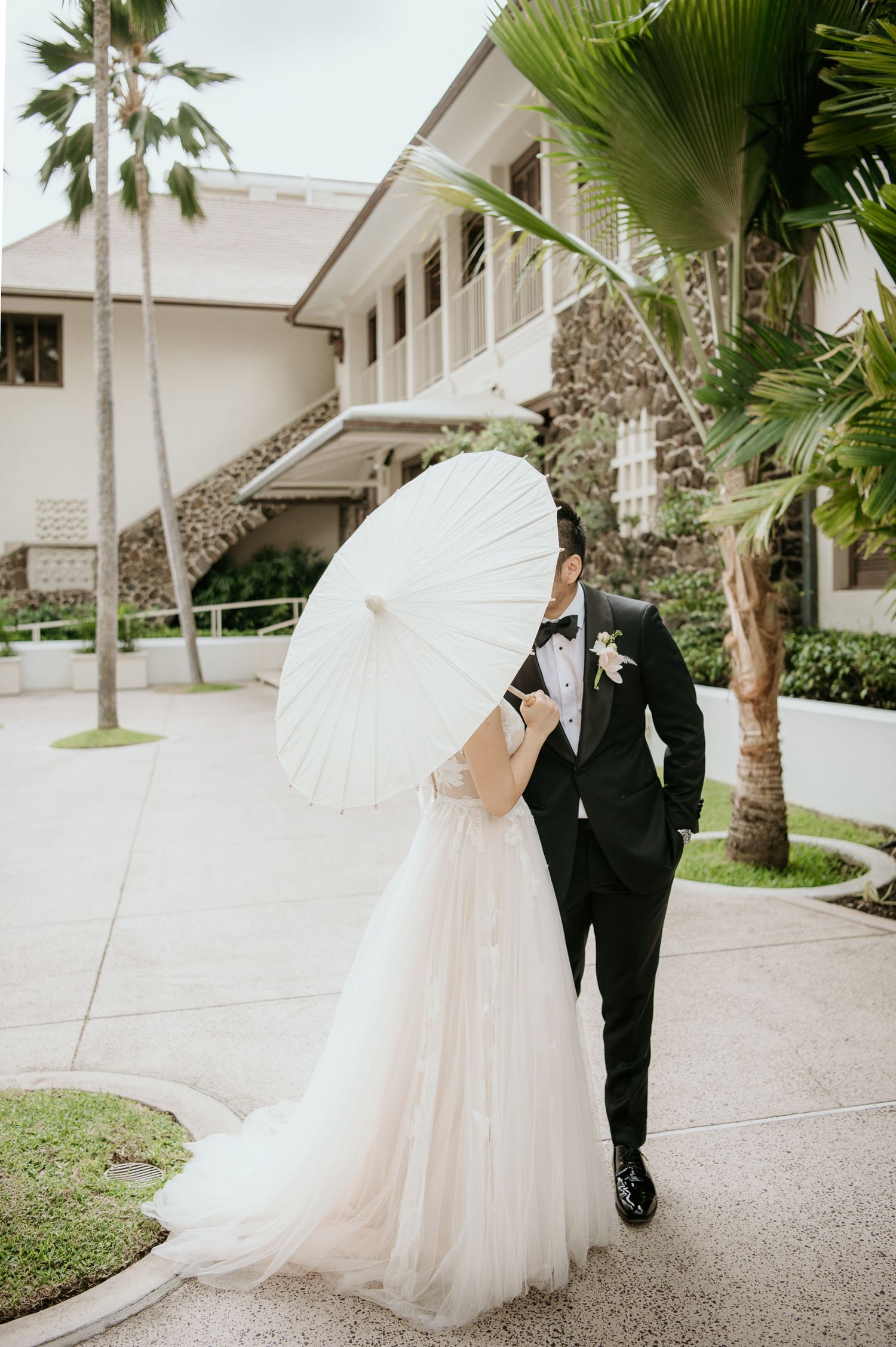 A romantic wedding couple poses on a tropical path with a white parasol and palm trees at a stone and white building.