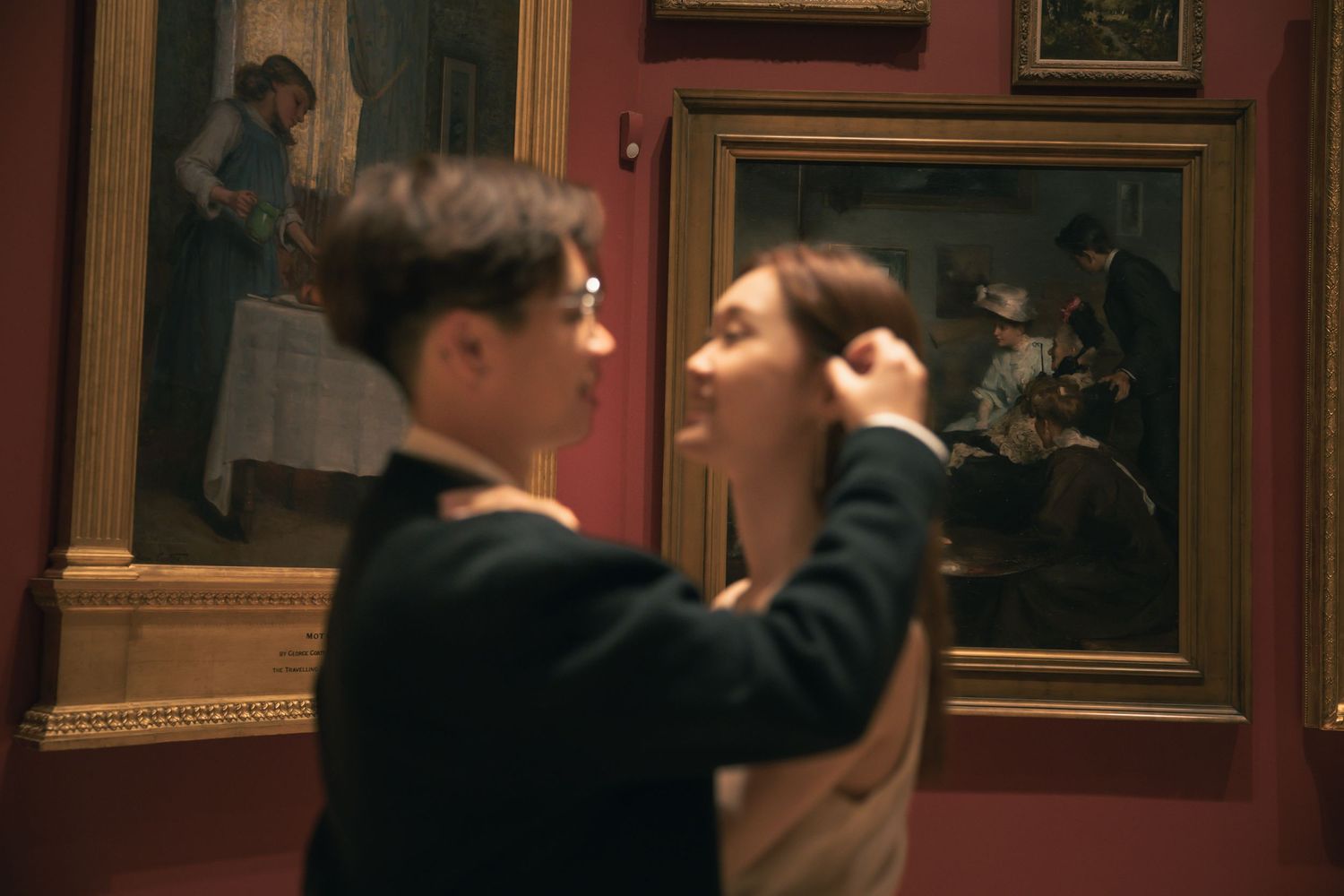 A romantic scene unfolds between two people in a museum gallery with red walls and ornate paintings.
