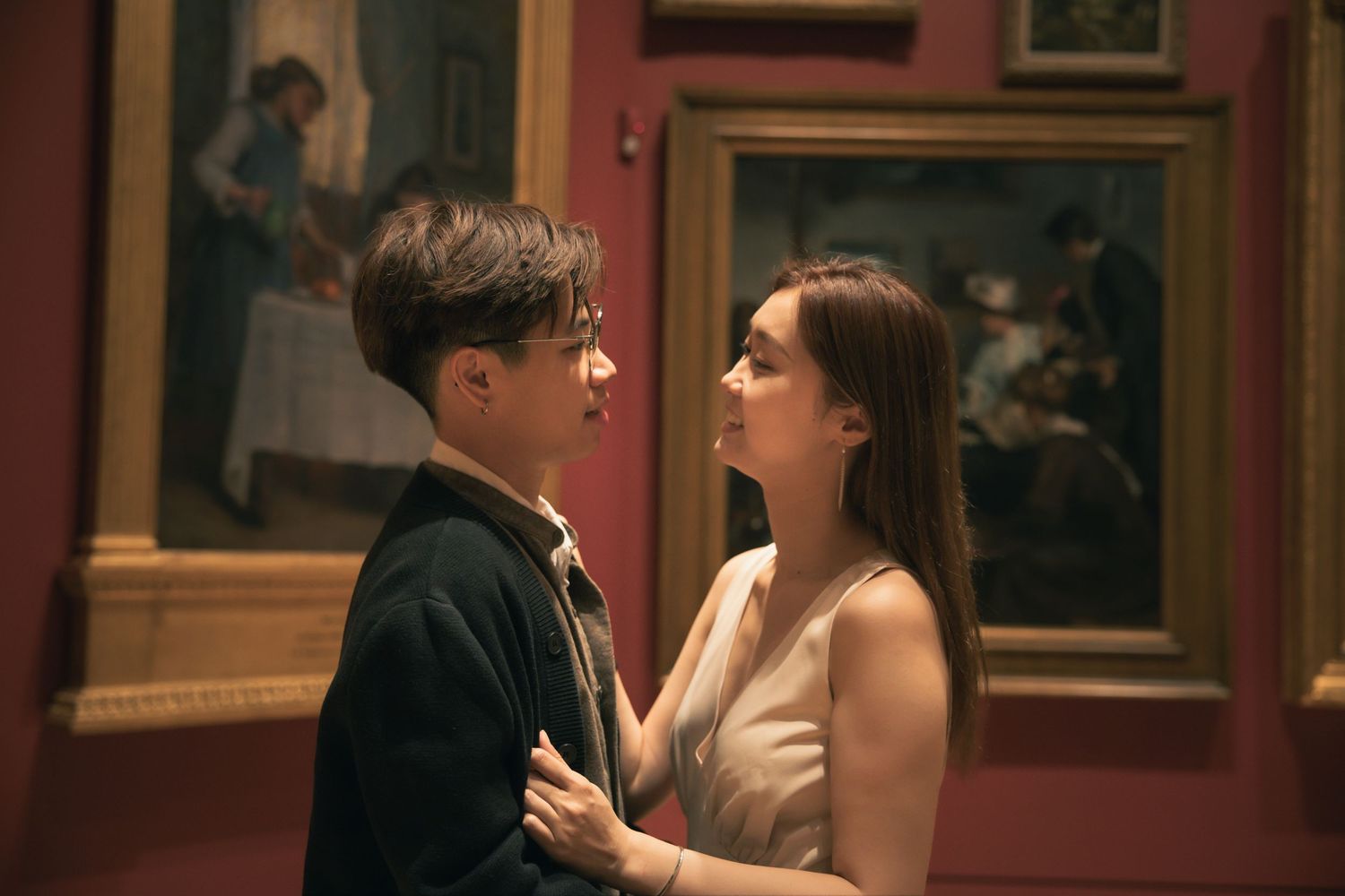 Two people share a tender moment in an art gallery with classical paintings on burgundy walls.
