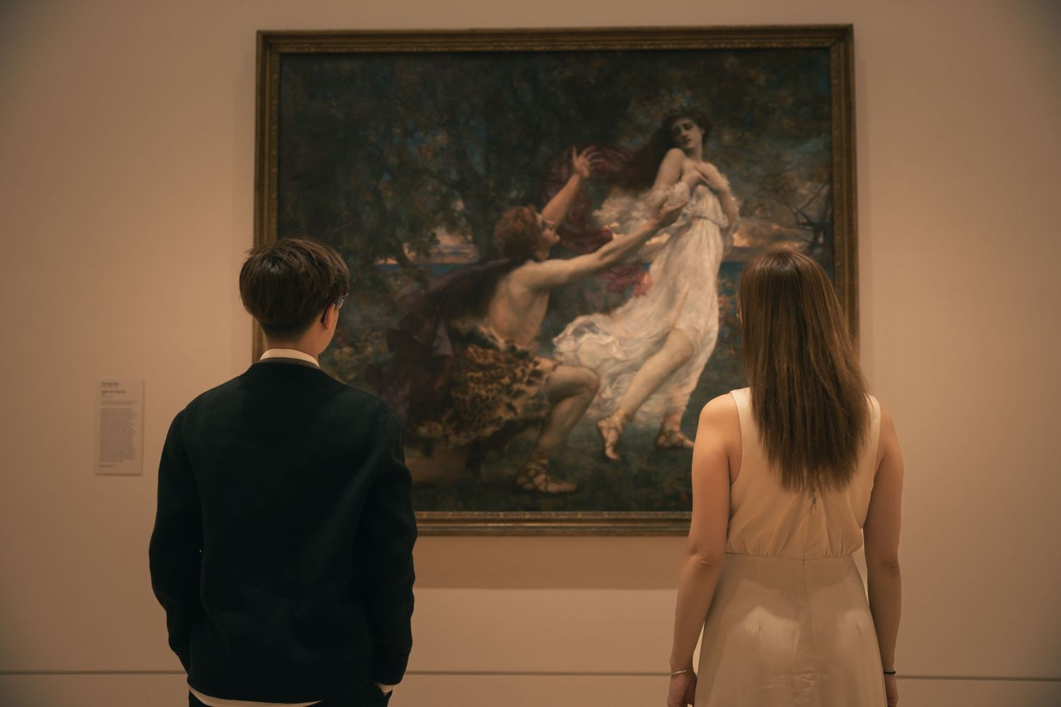 Two people admire a classical painting depicting a ethereal scene in a museum.