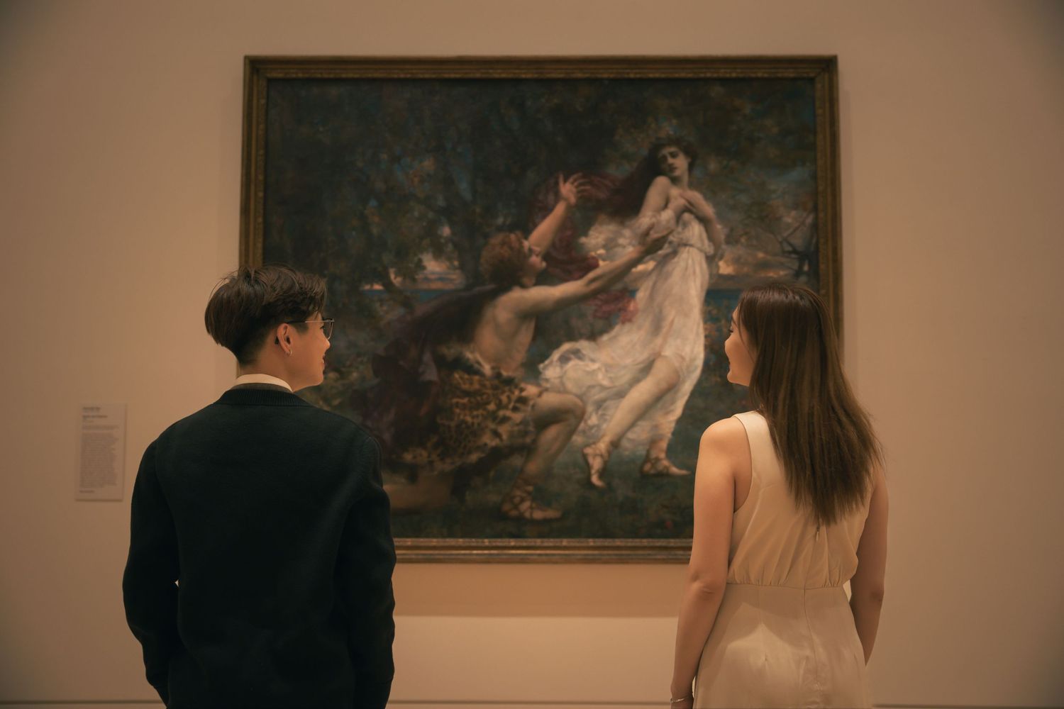 A couple views a dramatic classical painting in a dimly lit museum gallery.