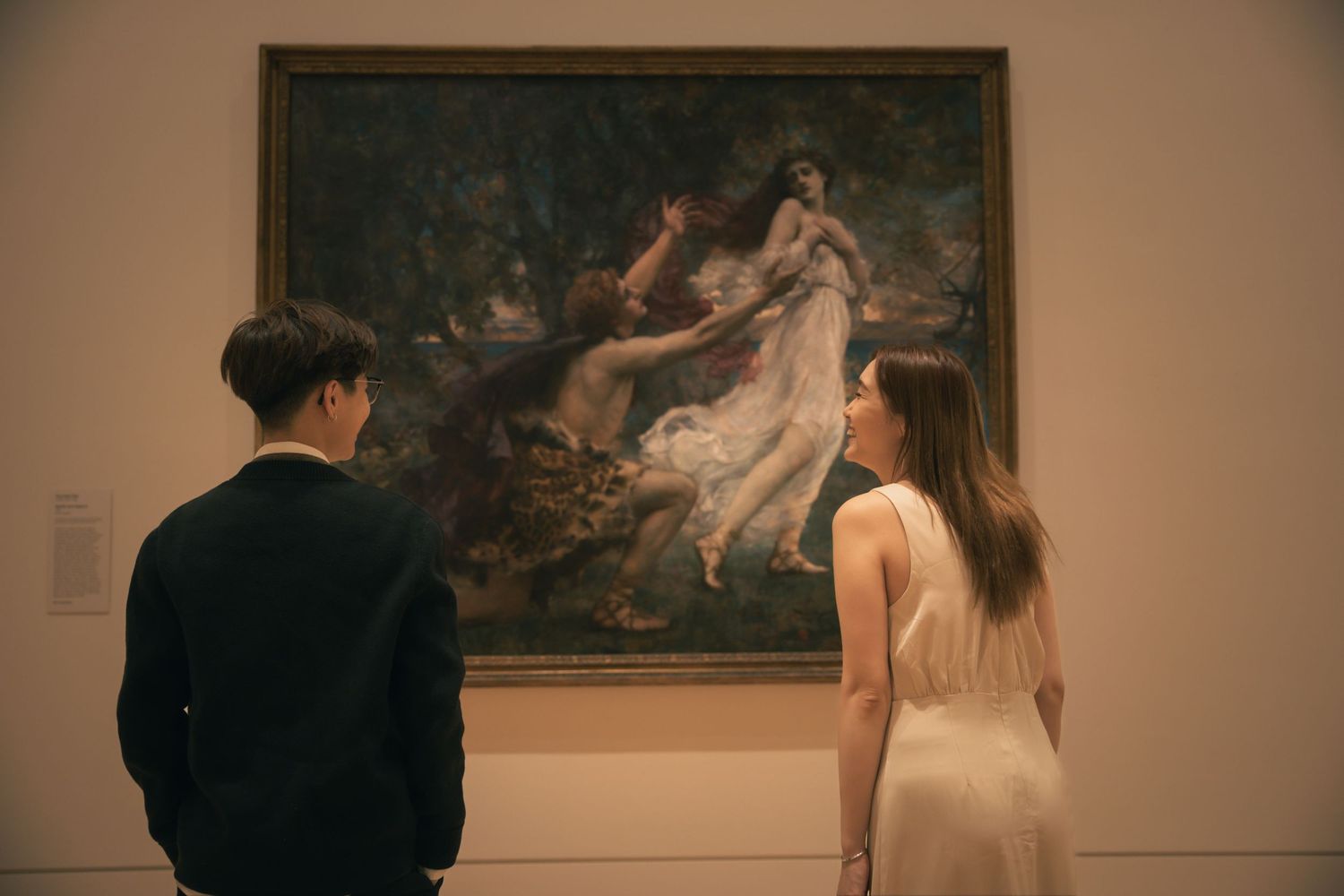 Two people observe a romantic classical painting featuring flowing figures in an art museum.