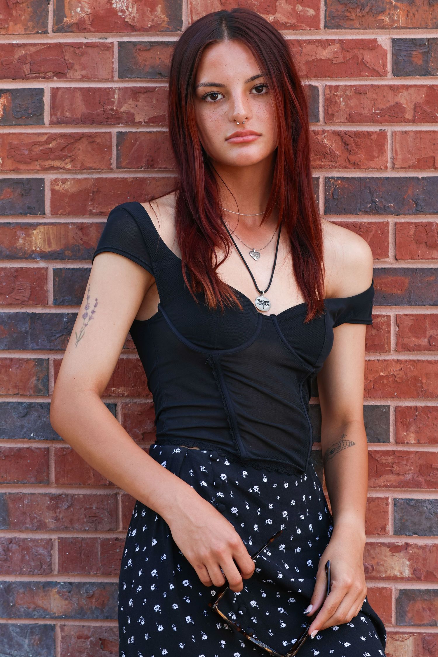 Woman in black off-shoulder top and patterned skirt poses against red brick wall outdoors for her senior portrait session.
