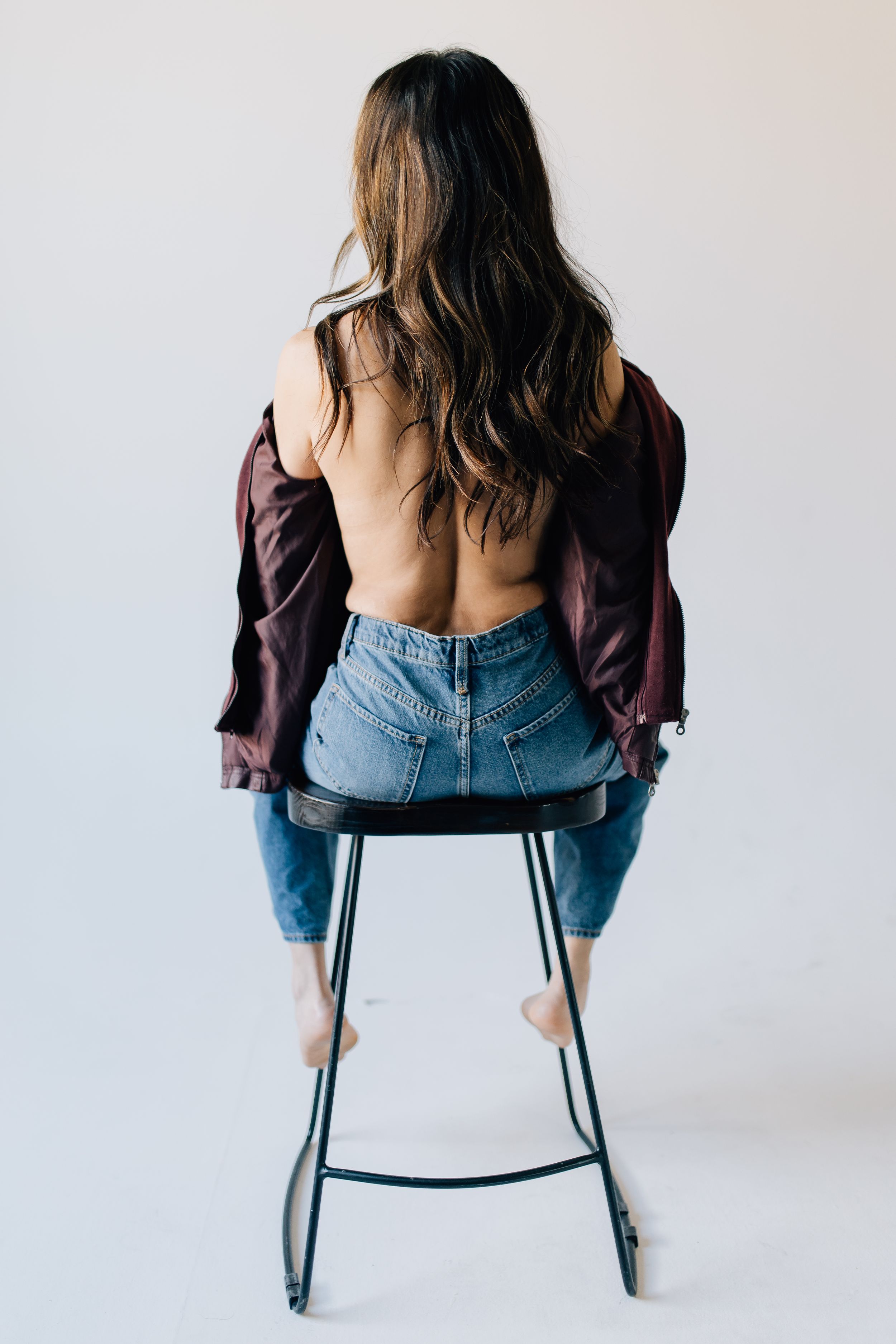 Woman in burgundy top and jeans sitting backwards on stool showing artistic backlit pose.
