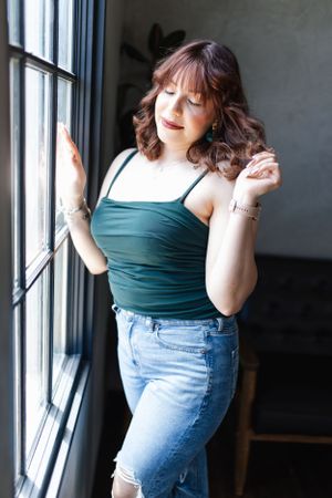 Woman in teal tank top and jeans poses by window in natural light.