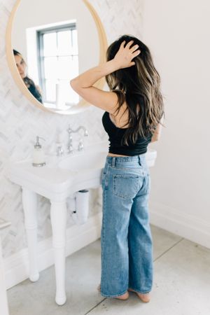 Woman in casual denim fixing her hair in front of round bathroom mirror.