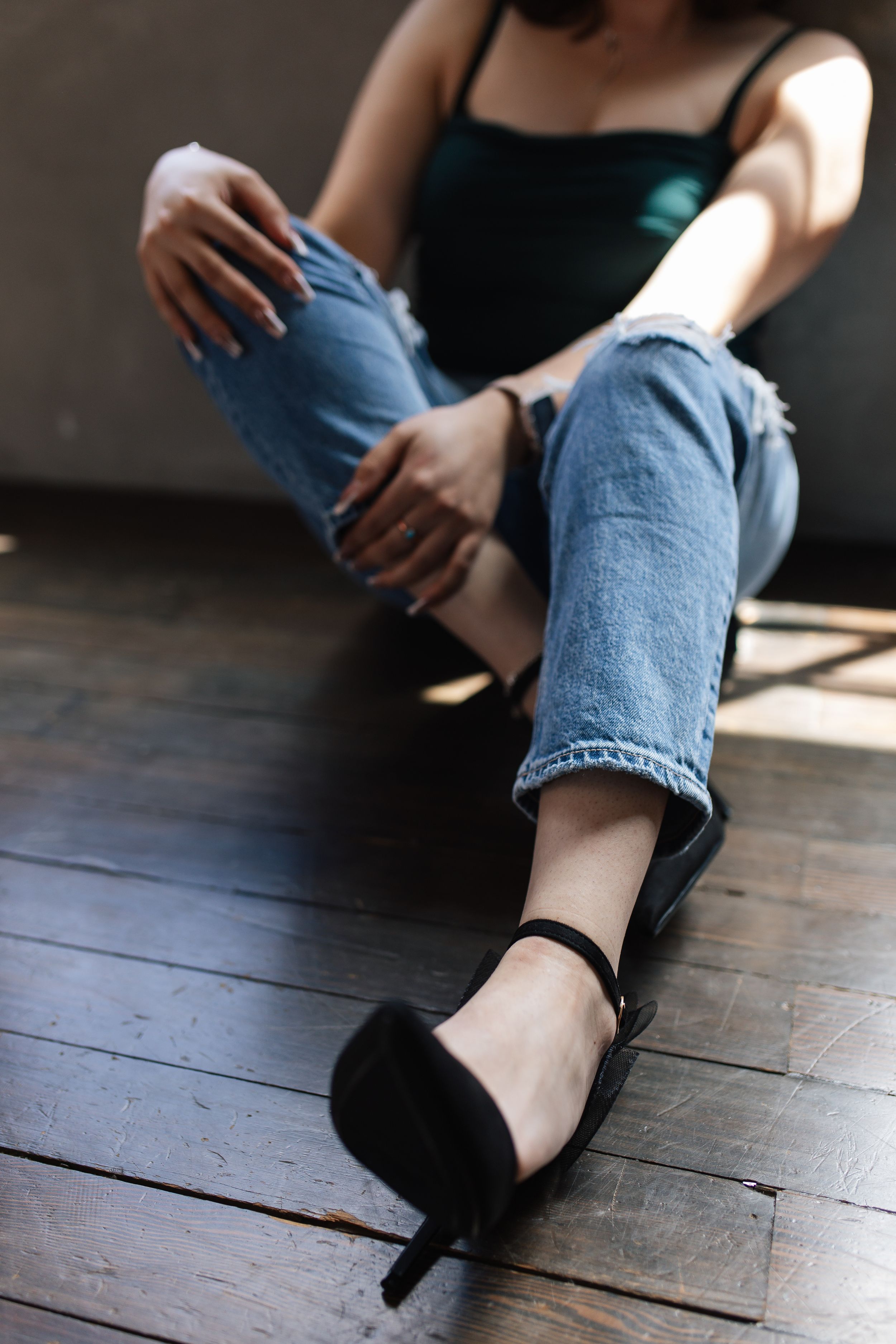 Person in distressed jeans and black heels sitting on wooden floor in moody lighting.