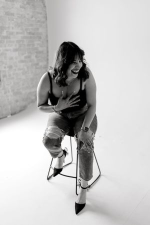 Black and white portrait of person laughing while sitting on stool in distressed jeans.