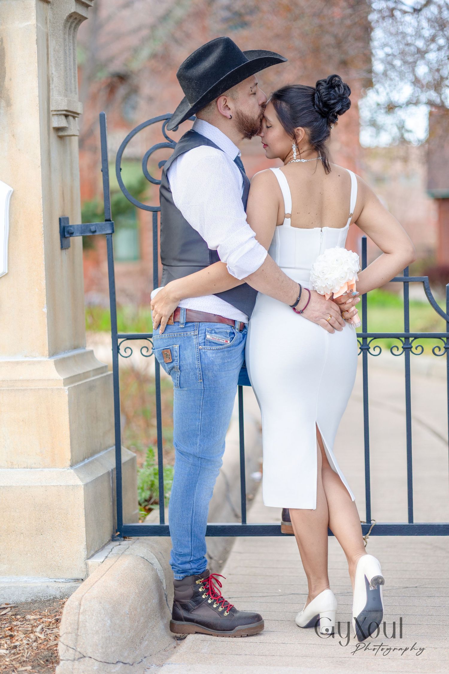 Romantic couple embraces by an ornate iron gate with the person in a cowboy hat and white shirt leaning against stone pillar.