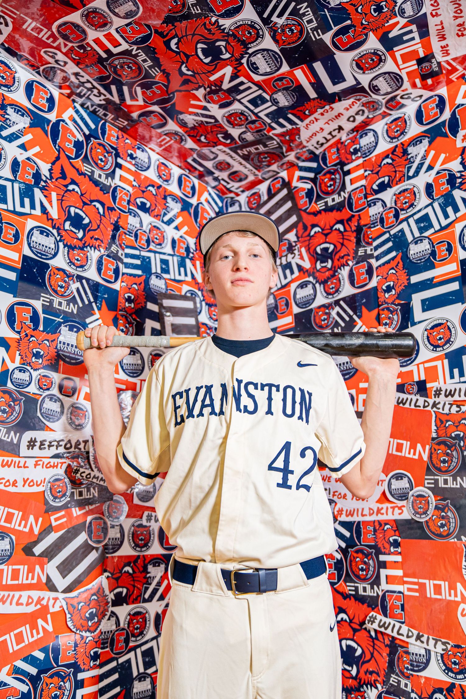 Baseball player stands holding bat in front of wall covered with red and blue Illinois team logos and pennants.