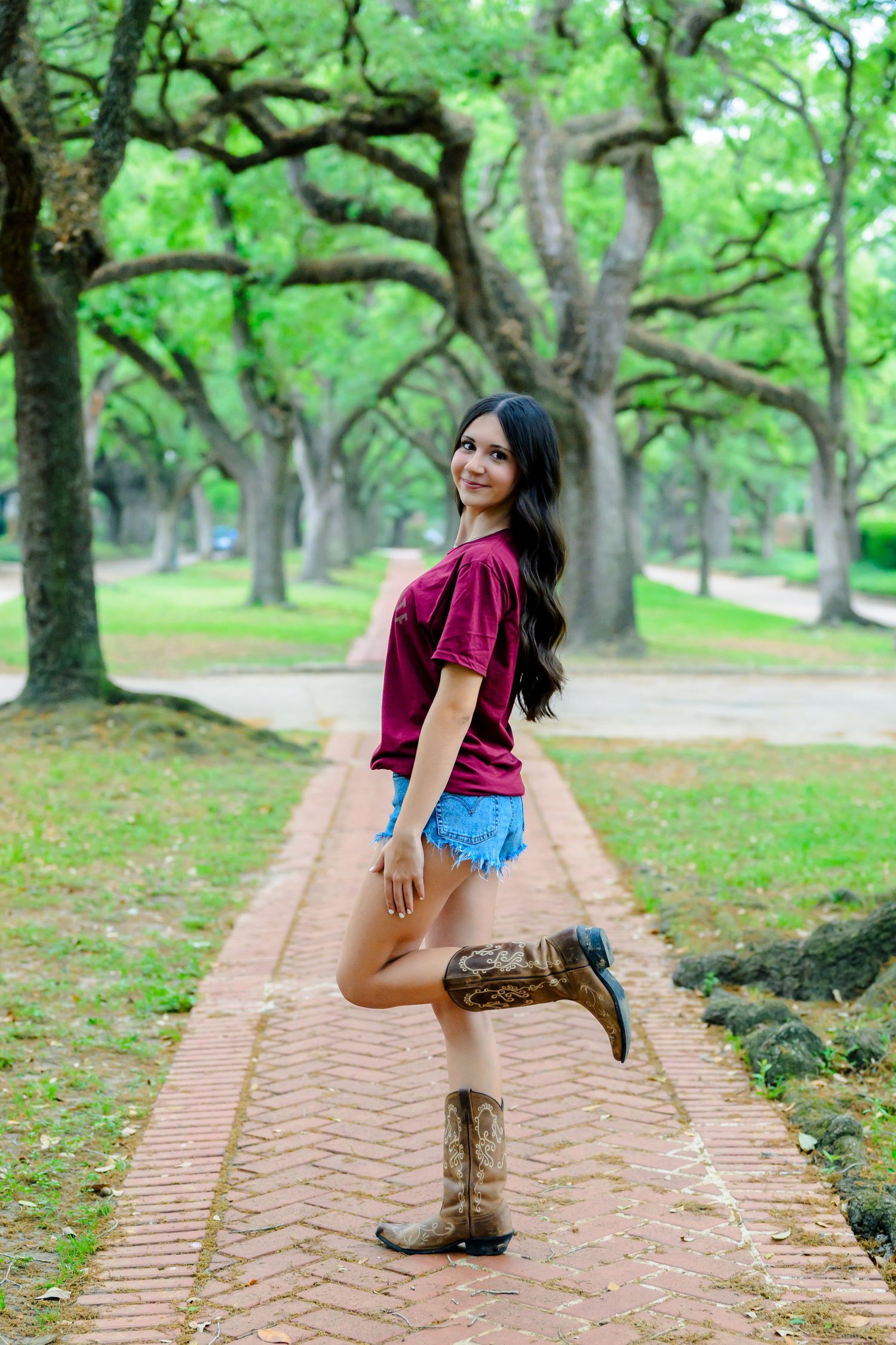 Someone poses playfully on a brick path surrounded by old oak trees wearing a burgundy top, denim shorts and cowboy boots.