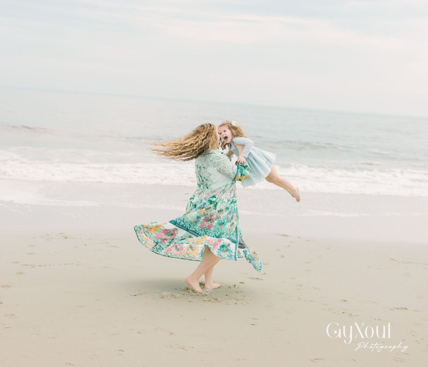 A person in a floral mint dress twirls joyfully on a misty beach with their hair flowing in the wind.