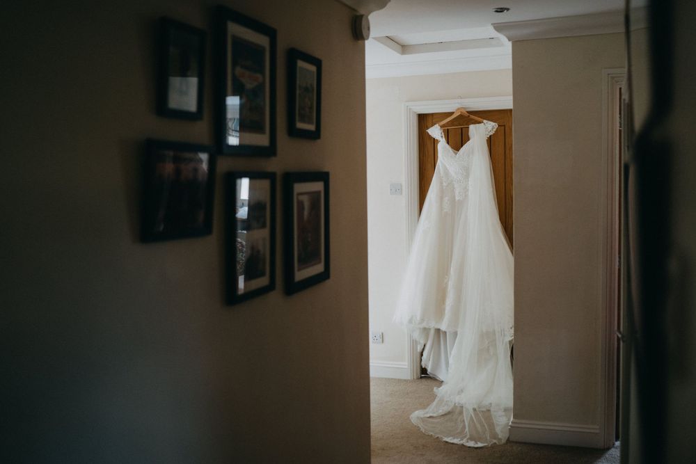 A white wedding dress hangs elegantly in a doorway with framed photos on the wall in the foreground.