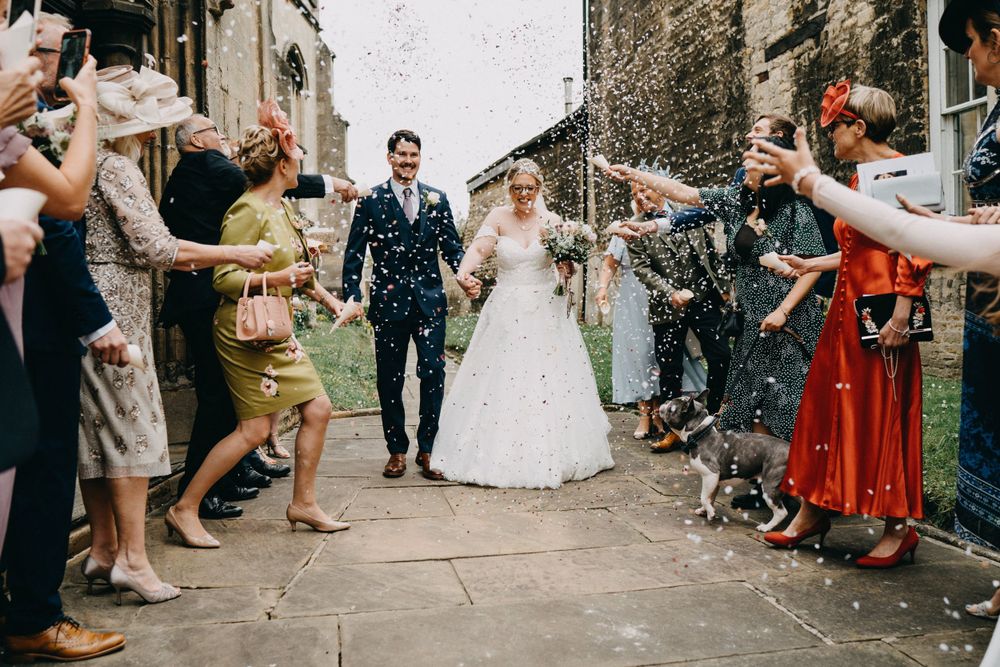 Wedding guests throw confetti over newlyweds during a joyful celebration outside a stone church building.