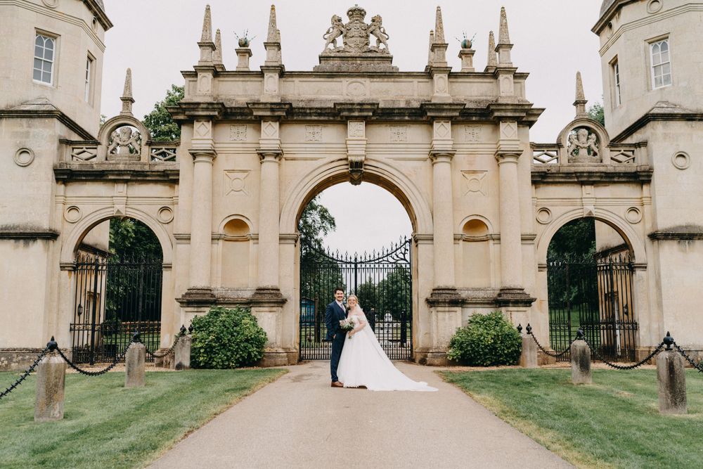 A couple stands in the ornate archway entrance of a grand historic estate with stone spires and decorative architecture.