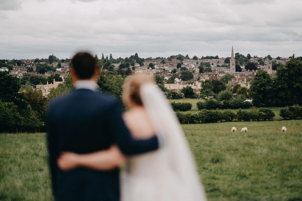 Newlyweds overlooking a pastoral English countryside landscape with village buildings and sheep grazing in green fields.