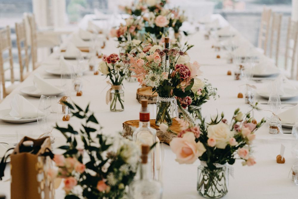 Elegant table setting with mason jar floral centerpieces featuring peach and pink roses along white linen tablecloth.