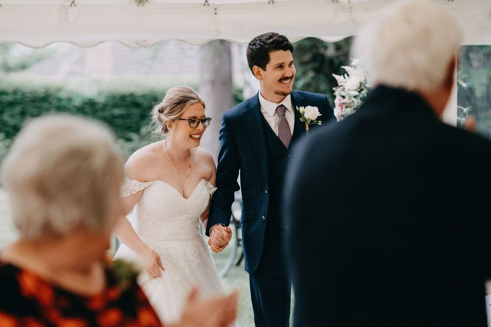 Newlyweds hold hands during outdoor wedding ceremony while guests look on from foreground.