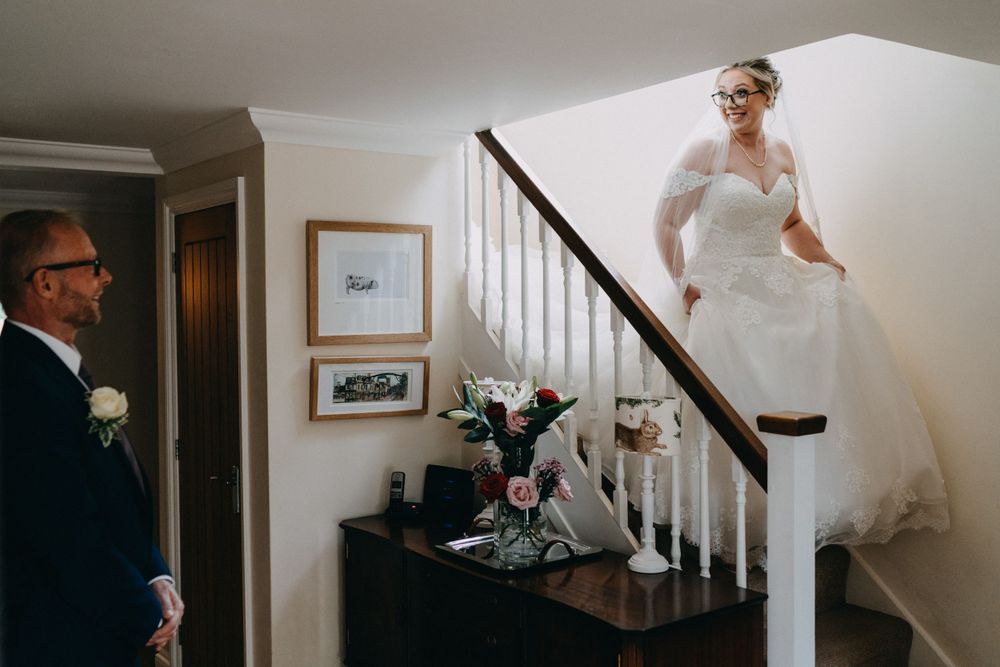 A person in a flowing white wedding dress descends a staircase while someone waits near a floral arrangement below.