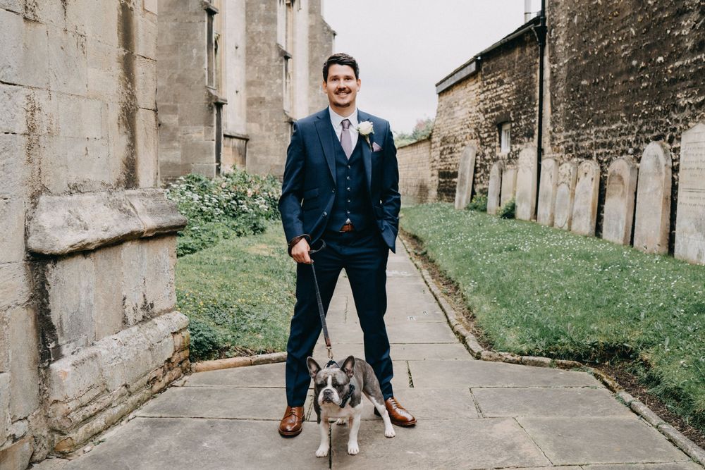 A groom in a navy suit stands with a small gray and white dog on a path between stone church walls.