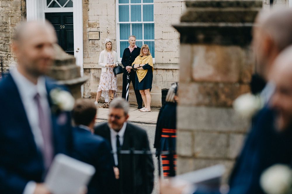 A candid wedding photo taken outside a stone building with guests and wedding party members in formal attire.