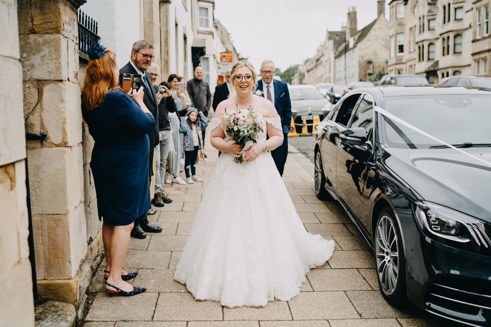 A joyful scene outside with onlookers waving at a person in an elegant white wedding gown next to a black luxury car.