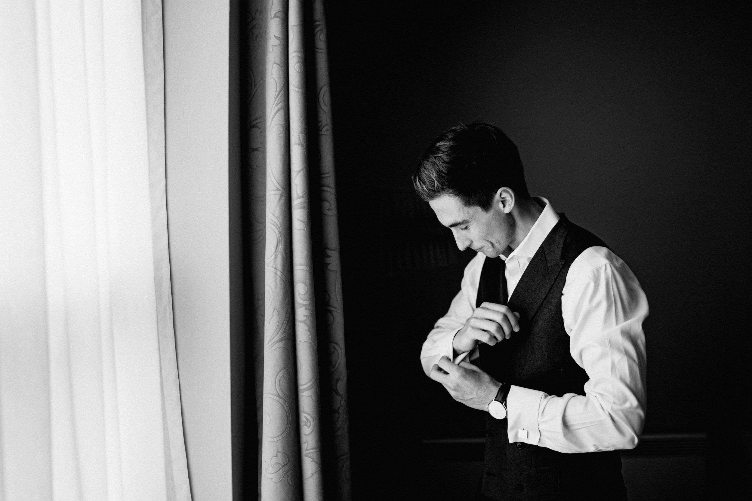 Black and white portrait of person in formal vest and tie adjusting cufflinks near window.