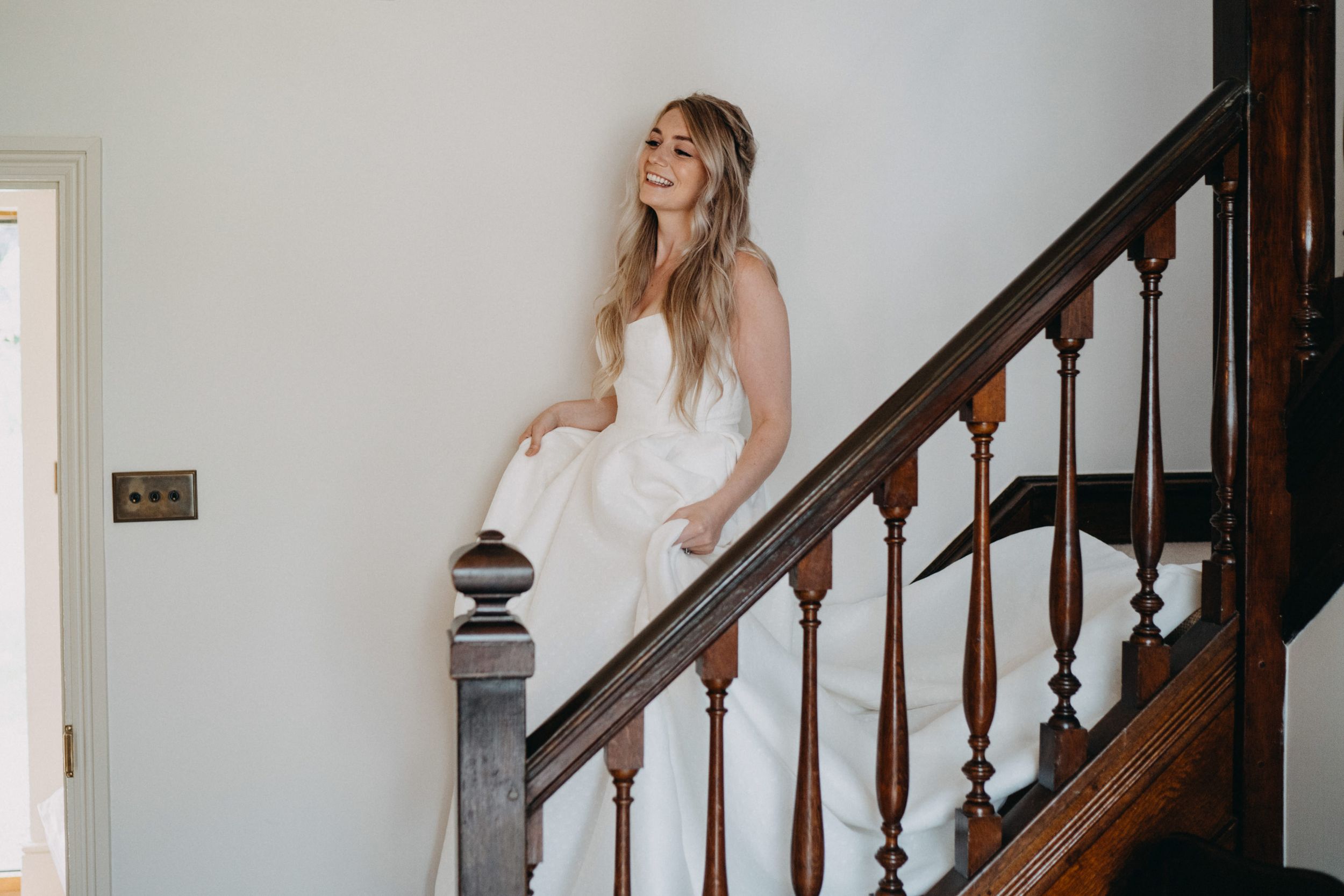 A bride in a white dress stands elegantly on a wooden staircase with ornate dark bannisters in a bright interior setting.