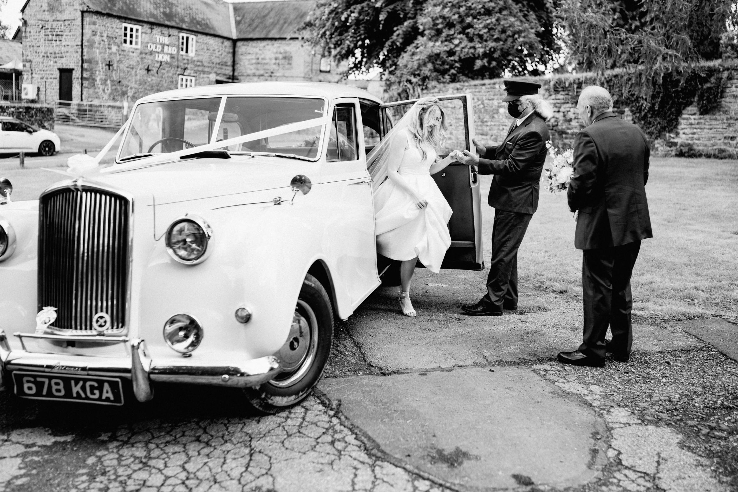 A classic white Bentley wedding car parked outside a stone building with passengers entering the vehicle.
