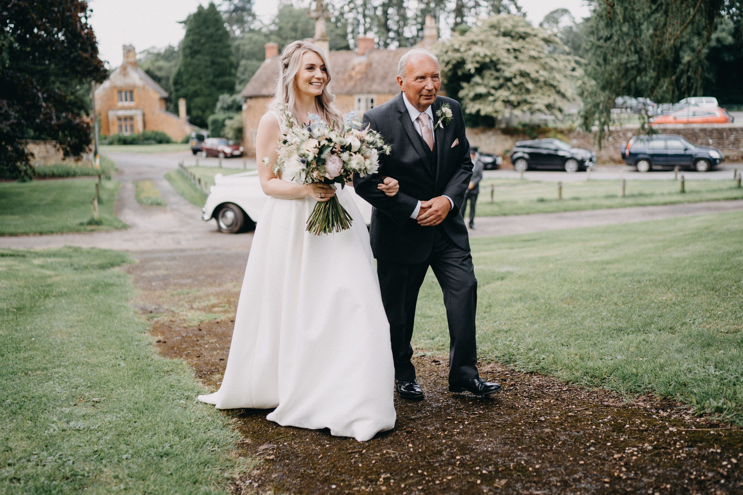 A person in a stunning white wedding dress with a bouquet walks arm in arm with someone in a black suit on a path.