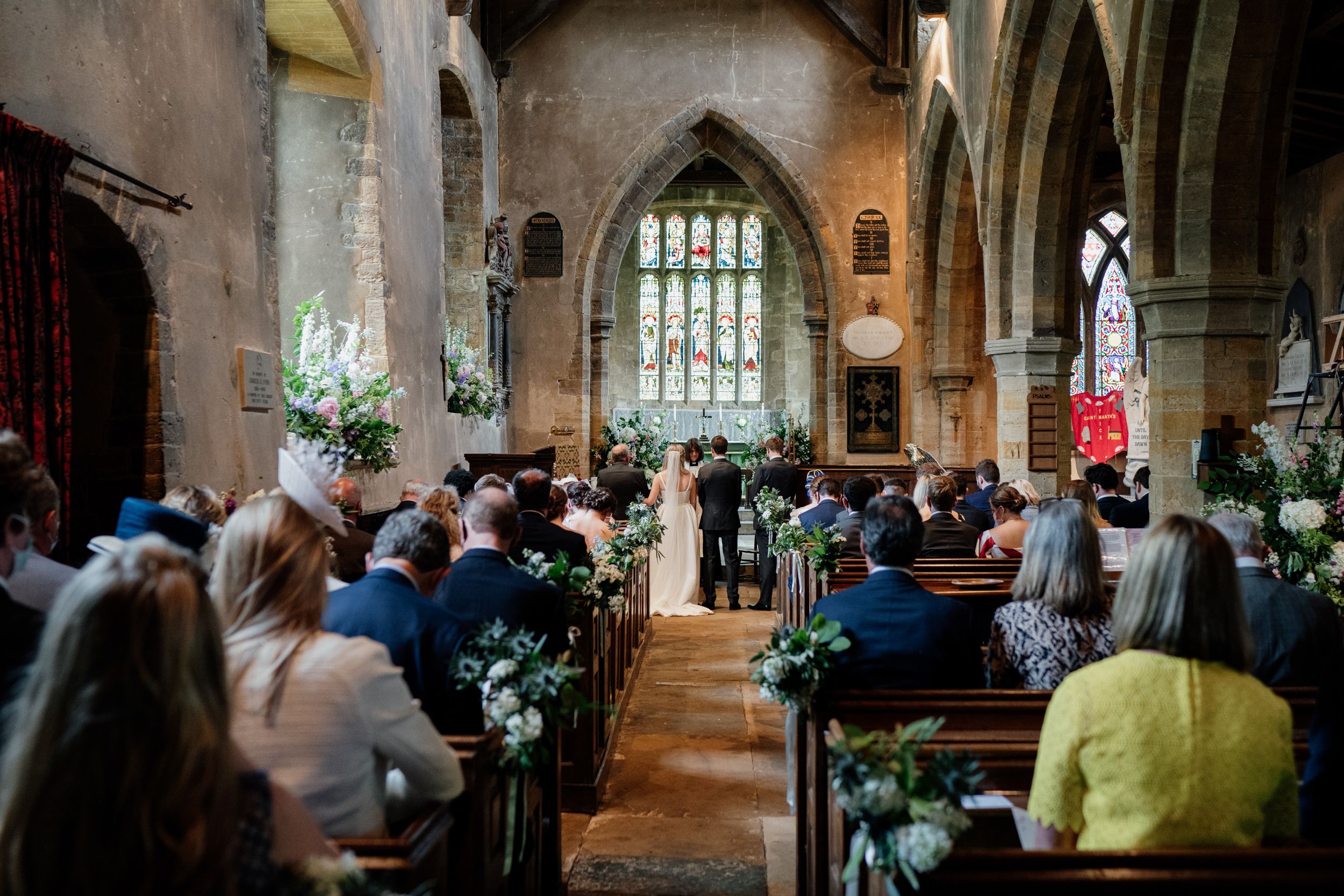 Wedding ceremony taking place in historic stone church with stained glass windows and elegant floral decorations.