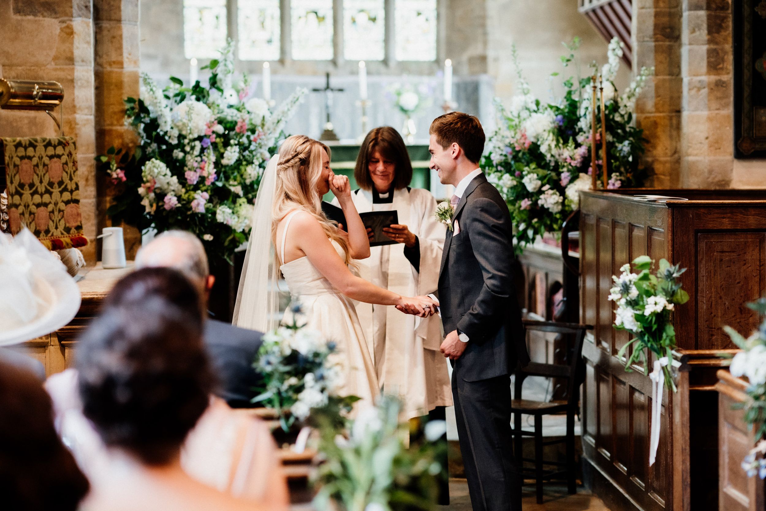 A couple stands at the altar during their wedding ceremony in a church adorned with elegant green and white floral arrangements.