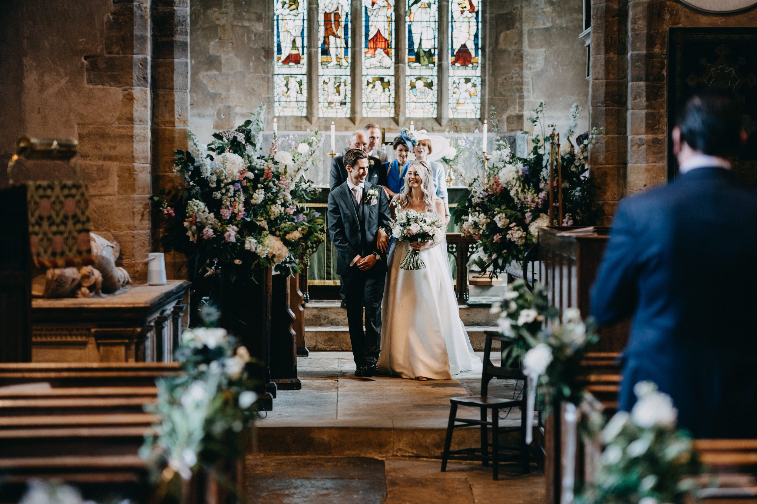 A wedding ceremony takes place in a historic stone church with stained glass windows and floral decorations along the aisle.