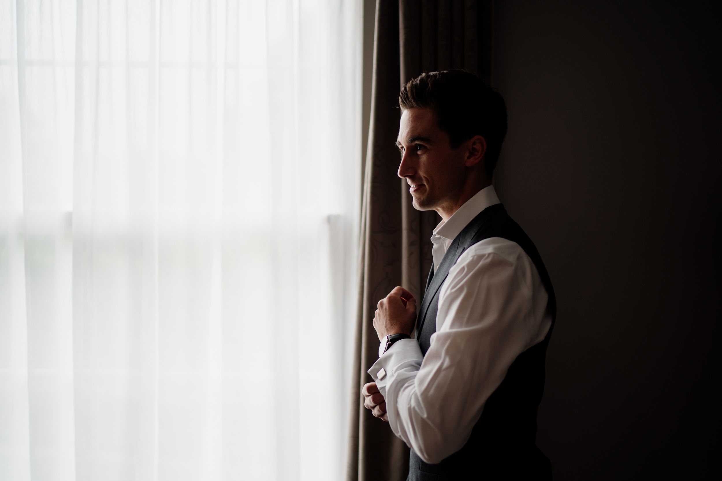 Silhouetted figure in white dress shirt stands contemplatively by window with sheer curtains in moody lighting.