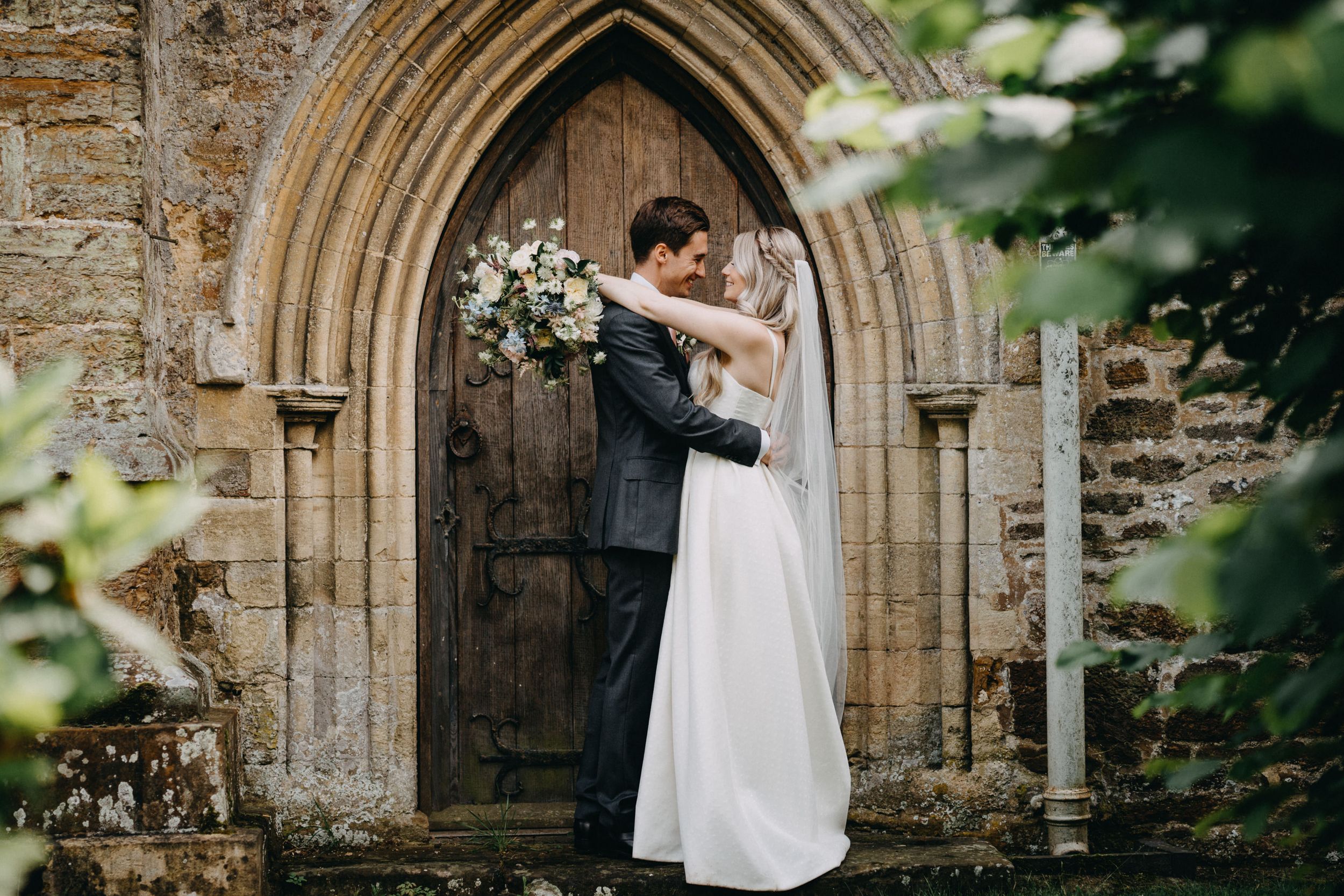 A romantic wedding couple embraces in front of a rustic stone church archway adorned with floral decorations.