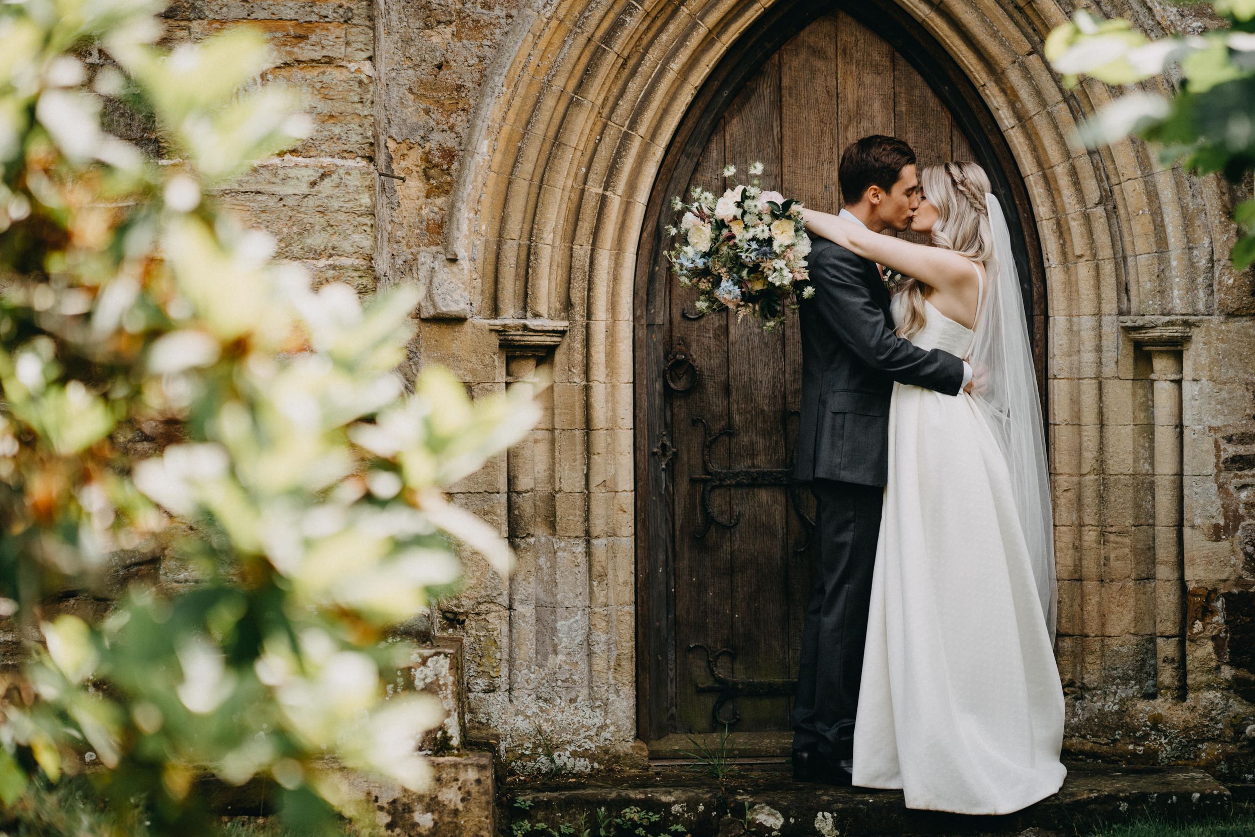 A couple shares a romantic embrace in front of an ornate stone church doorway with gothic arches and floral arrangements.