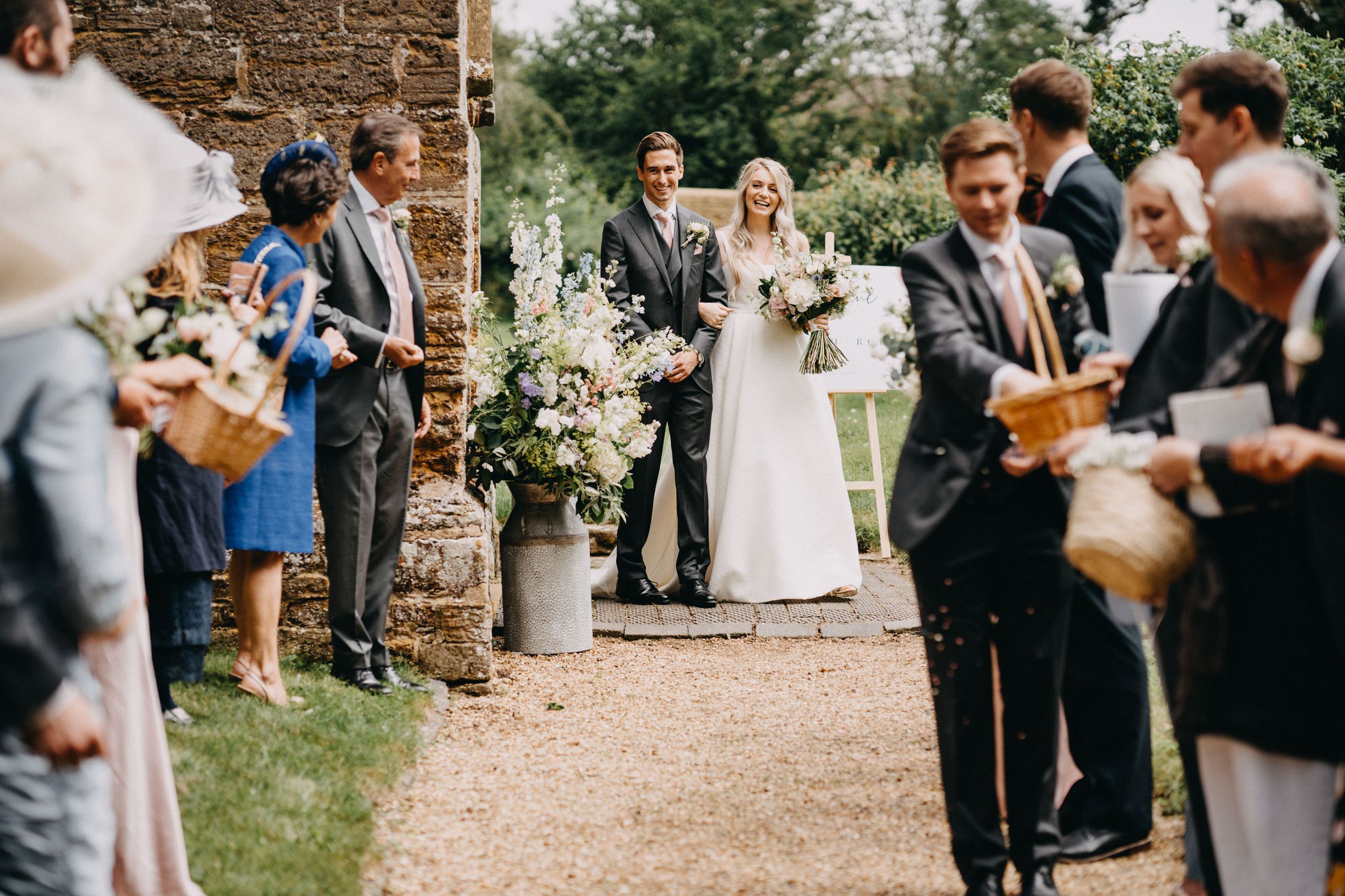 Wedding guests hold wooden photo frames as a couple stands on a rustic stone pathway during an outdoor ceremony.