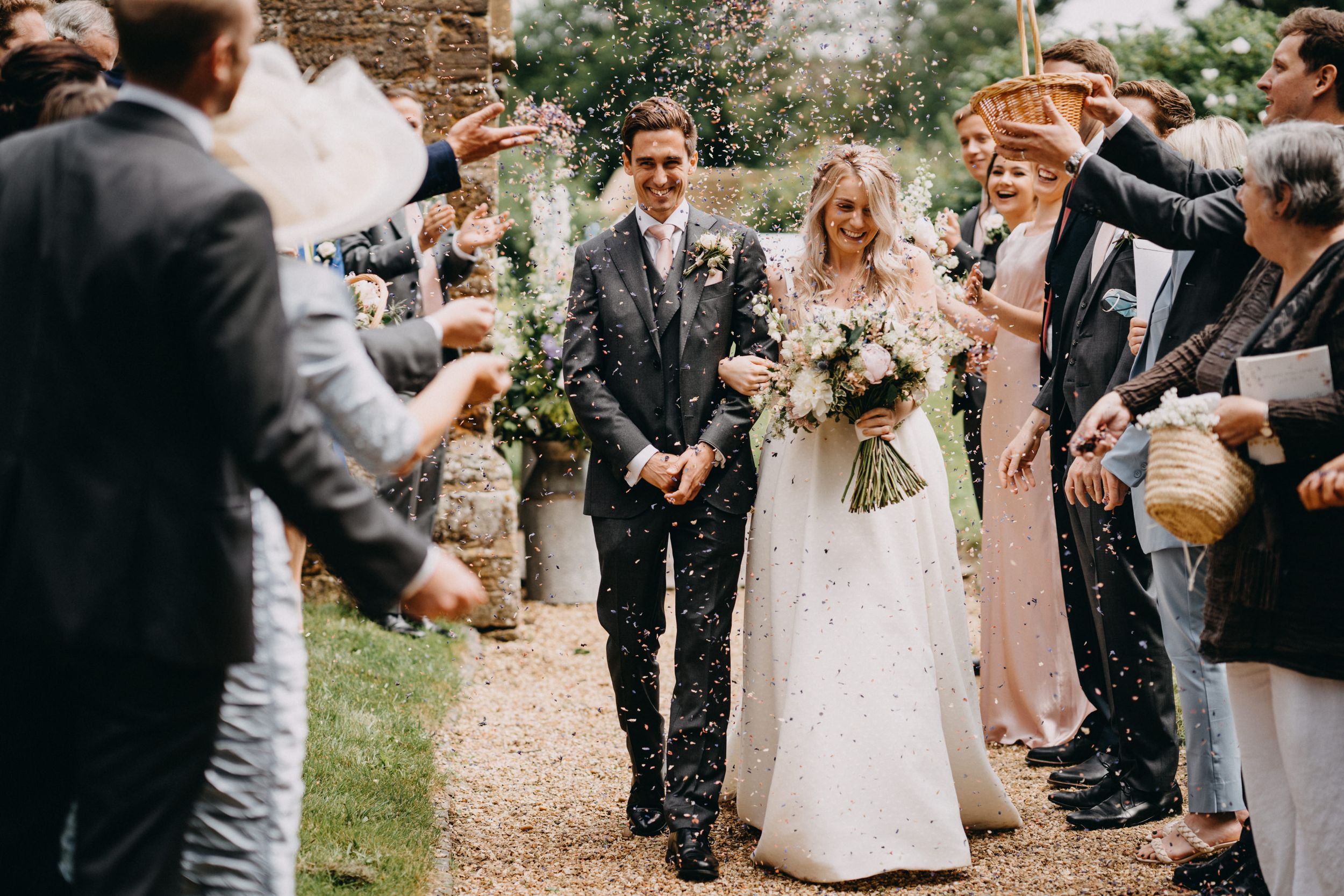 Wedding guests throw confetti over newlyweds walking down a garden path after their ceremony.